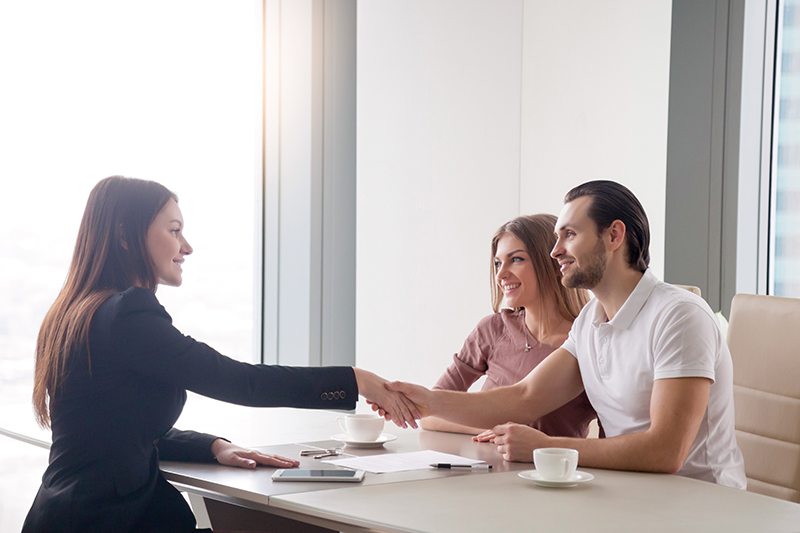 Young prosperous couple meeting with female broker in real estate agency. Man and real estate agent shaking hands, happy to make a deal of buying property, investment advisor congratulating clients