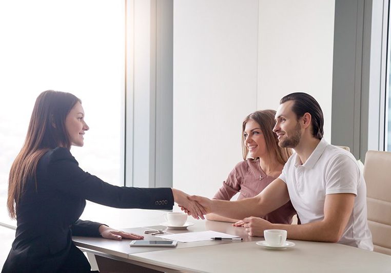 Young prosperous couple meeting with female broker in real estate agency. Man and real estate agent shaking hands, happy to make a deal of buying property, investment advisor congratulating clients